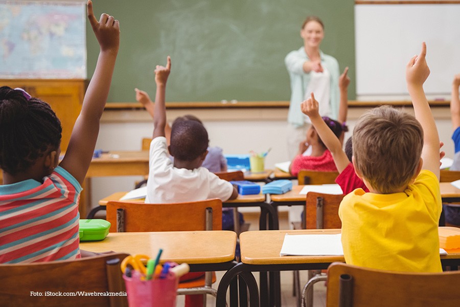 KI generiert: Das Bild zeigt eine Lehrerin vor einer Klasse von Schülern, die ihre Hände heben, um sich zu melden. Die Szene spielt sich in einem Klassenzimmer ab, mit einer Tafel im Hintergrund.