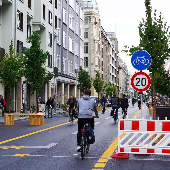 KI generiert: Das Bild zeigt eine belebte städtische Straße, die hauptsächlich von Fahrradfahrern genutzt wird. Schilder deuten darauf hin, dass es sich um eine verkehrsberuhigte Zone mit einer Geschwindigkeitsbegrenzung von 20 km/h handelt.