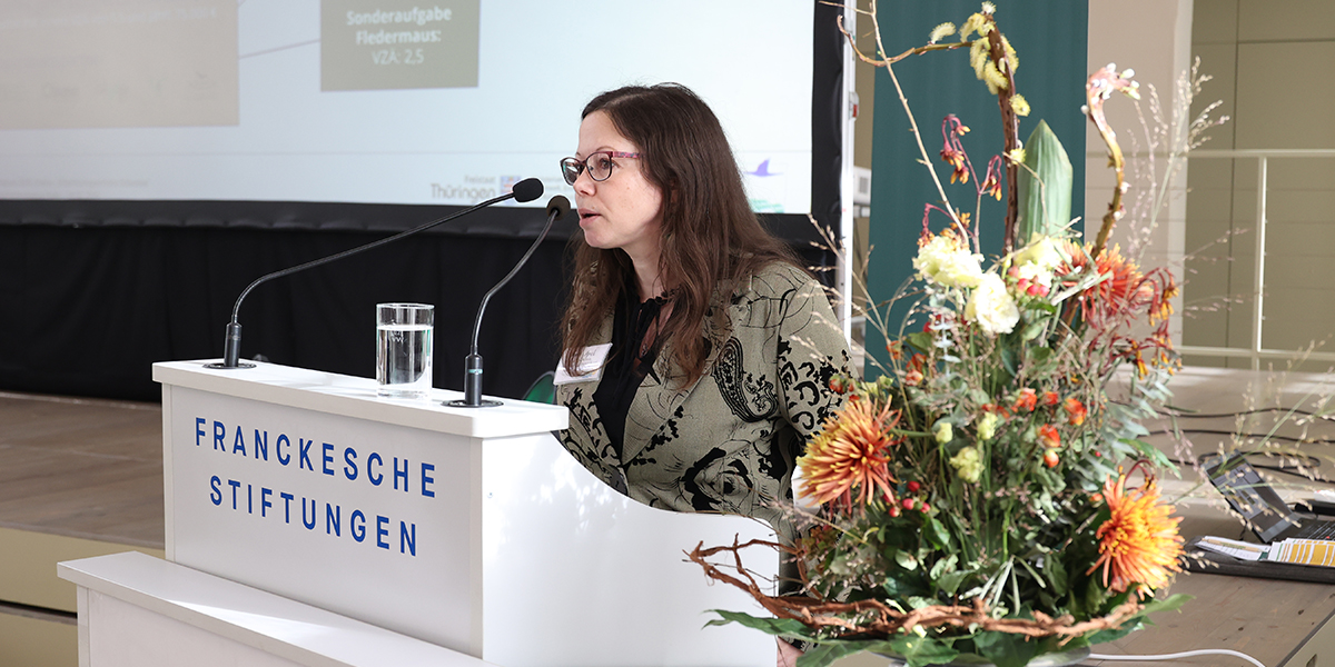 KI generiert: Eine Frau hält einen Vortrag an einem Rednerpult der Franckeschen Stiftungen, während rechts davon ein Blumenarrangement zu sehen ist.