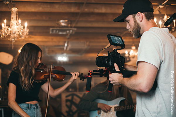 KI generiert: Das Bild zeigt einen Filmemacher, der eine Musikerin mit einer Violine in einem Innenraum filmt, der von Kronleuchtern beleuchtet wird. Im Hintergrund ist eine weitere Person mit einer Gitarre zu sehen.