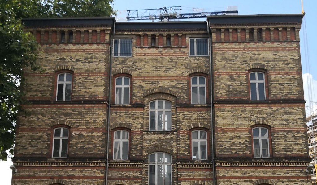 KI generiert: Das Bild zeigt die Fassade eines älteren Backsteingebäudes mit symmetrisch angeordneten Fenstern. Auf dem Dach ist ein Baukran sichtbar, was auf Bauarbeiten oder Renovierungen hindeutet.