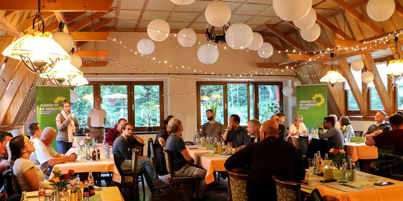 KI generiert: Das Bild zeigt ein Treffen von Menschen in einem dekorierten Raum mit Tischen, auf denen Getränke stehen. Die Anwesenden scheinen einer Person zuzuhören, die gerade spricht, und im Hintergrund sind Banner von Bündnis 90/Die Grünen zu sehen.