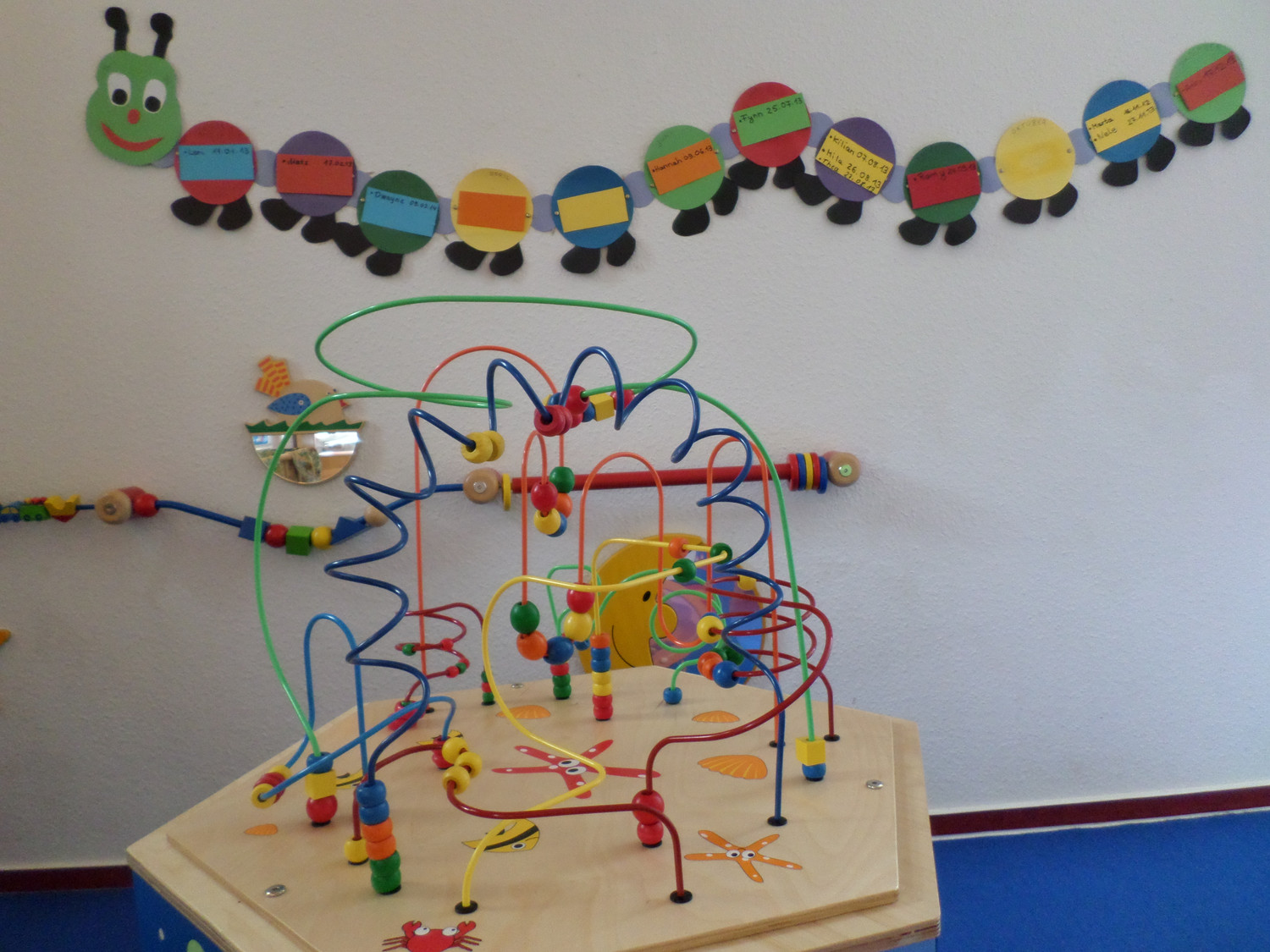 KI generiert: Das Bild zeigt ein buntes Motorikspielzeug mit verschlungenen Drähten und Holzkugeln, das auf einem Tisch steht. Im Hintergrund ist eine dekorative Raupe aus buntem Papier an der Wand zu sehen.