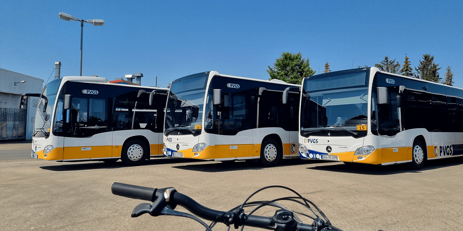 KI generiert: Das Bild zeigt drei geparkte Stadtbusse der PVGS auf einem Busdepot bei strahlend blauem Himmel. Im Vordergrund ist ein Teil eines Fahrradlenkers zu sehen.