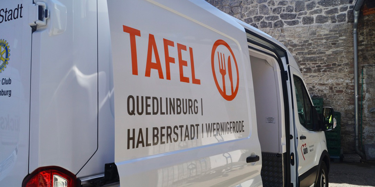 KI generiert: Das Bild zeigt einen weißen Lieferwagen mit der Aufschrift "TAFEL QUEDLINBURG | HALBERSTADT | WERNIGERODE" und dem Symbol eines Bestecksets. Der Transporter steht vor einer Steinmauer und symbolisiert die Mobilität und den Service der Tafel in diesen Städten.
