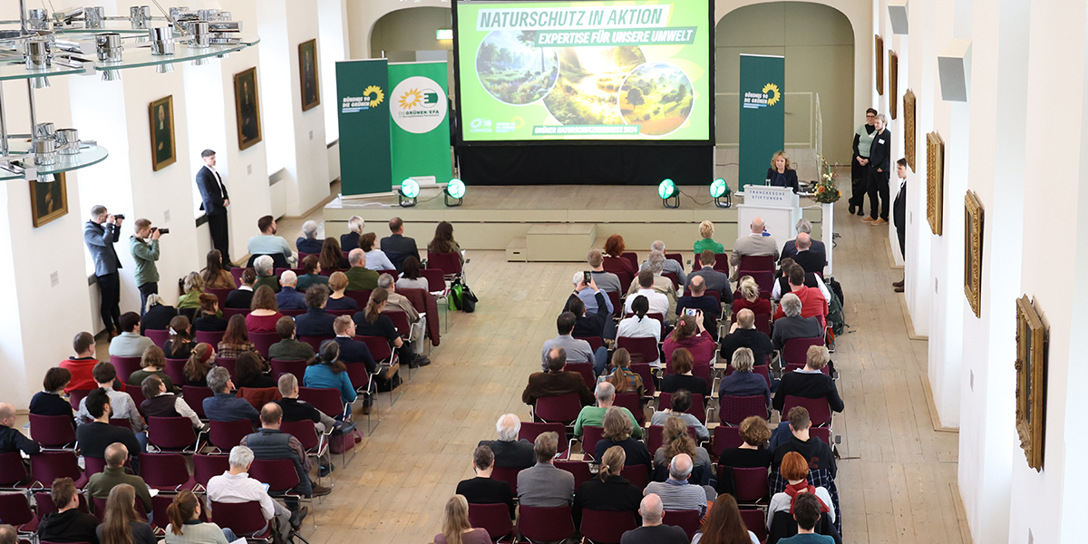 KI generiert: Das Bild zeigt eine Veranstaltung in einem Konferenzsaal, bei der eine Person am Podium zu einer sitzenden Menge spricht. Auf der Leinwand steht "Naturschutz in Aktion: Expertise für unsere Umwelt".