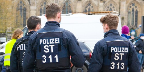 KI generiert: Das Bild zeigt mehrere Polizeibeamte in Uniform mit sichtbaren Rückenkennzeichnungen, die bei einer Veranstaltung oder einem Einsatz im Freien unterwegs sind. Im Hintergrund sind ein Transporter sowie weitere Personen erkennbar.
