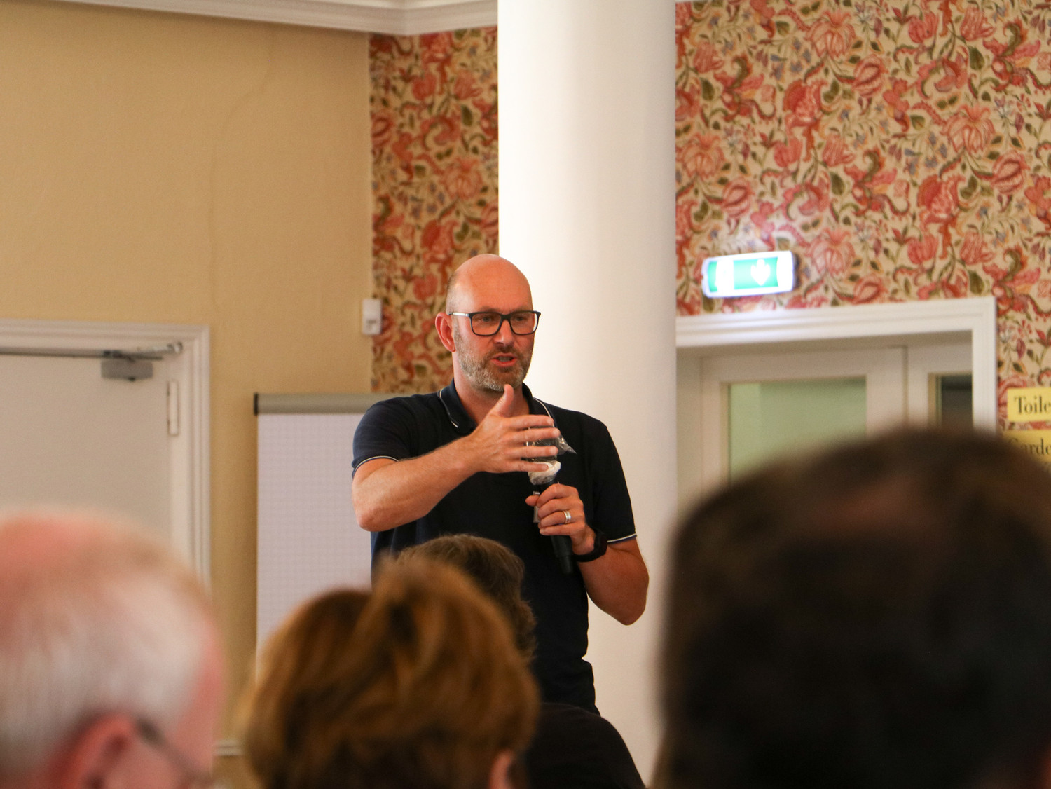 KI generiert: Ein Mann hält einen Vortrag vor einer Gruppe von Menschen, während er ein Mikrofon in der Hand hält. Er steht in einem Raum mit floral gemusterter Tapete und redet gestikulierend.