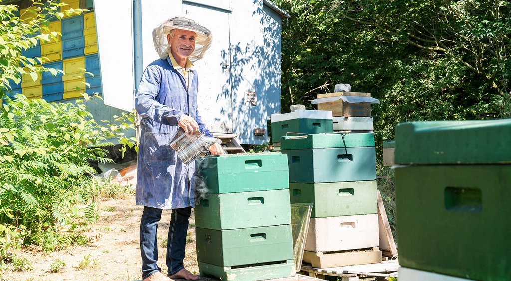 Enrico Kretschmar steht mit Imkerhut neben einem Bienenstock. Im Hintergrund befindet sich ein weiterer Wagen mit Bienenstöcken. 