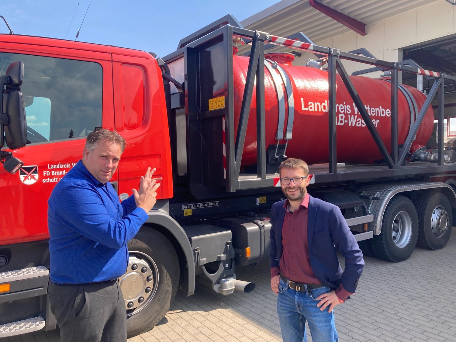KI generiert: Das Bild zeigt zwei Männer vor einem roten Feuerwehrfahrzeug mit einem großen Wassertank darauf. Sie posieren neben dem Fahrzeug, das anscheinend zu einer Feuerwehr im Landkreis gehört.