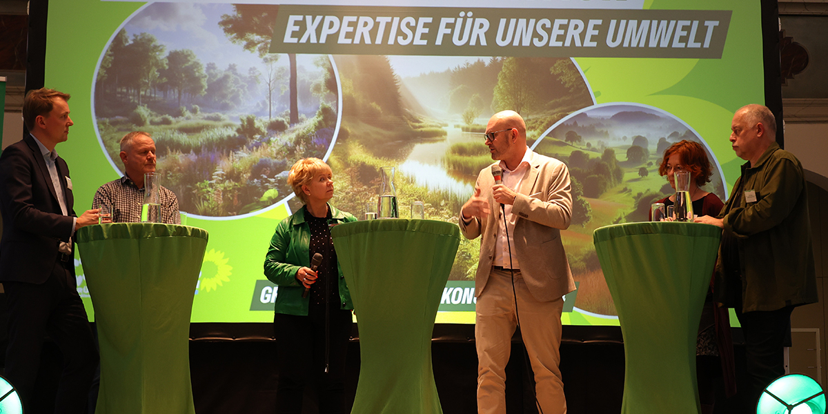 KI generiert: Das Bild zeigt eine Diskussionsrunde mit sechs Personen, die an Stehtischen vor einem Hintergrund mit der Aufschrift "Expertise für unsere Umwelt" stehen. Es handelt sich um eine Veranstaltung mit umweltbezogenem Thema.
