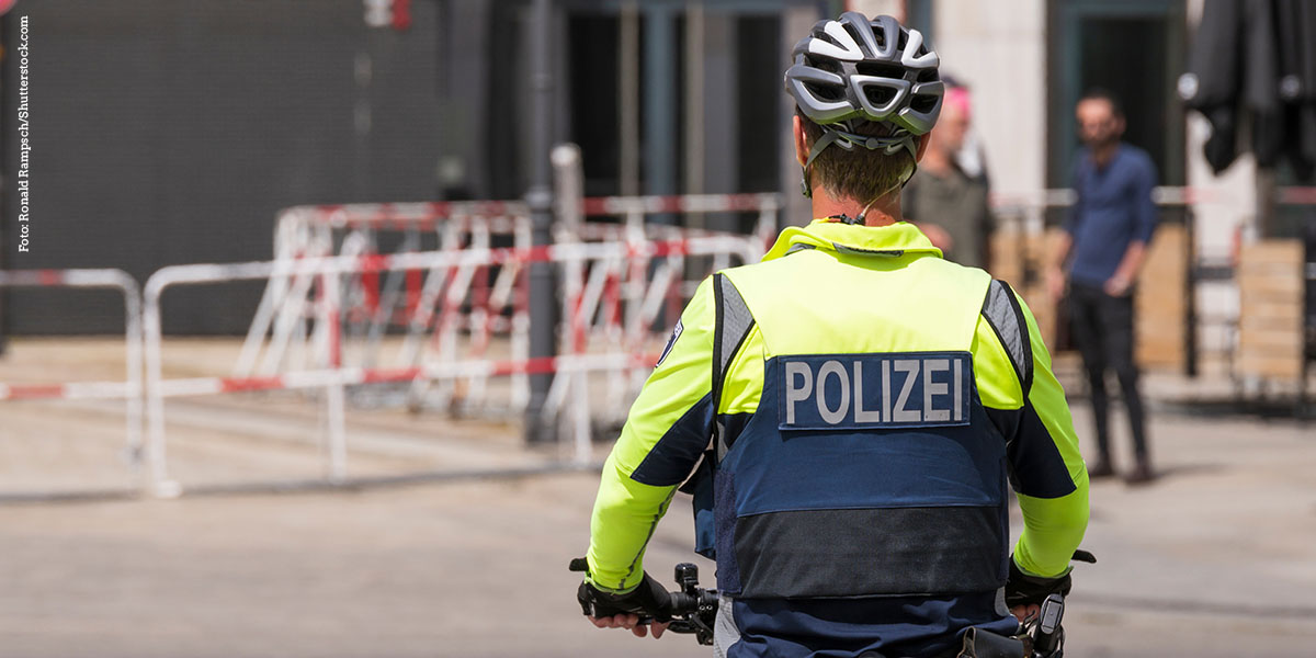 KI generiert: Das Bild zeigt einen Polizisten auf einem Fahrrad aus der Rückenansicht, bekleidet mit einer gelben Warnweste und einem Helm. Im Hintergrund sind Absperrungen und Gebäude zu sehen.