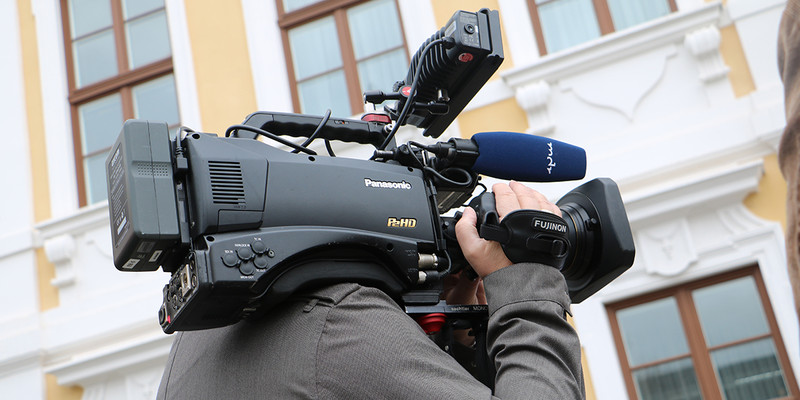 KI generiert: Ein Kameramann filmt mit einer großen professionellen Videokamera in einem städtischen Umfeld. Im Hintergrund sind Teile eines Gebäudes mit großen Fenstern zu sehen.