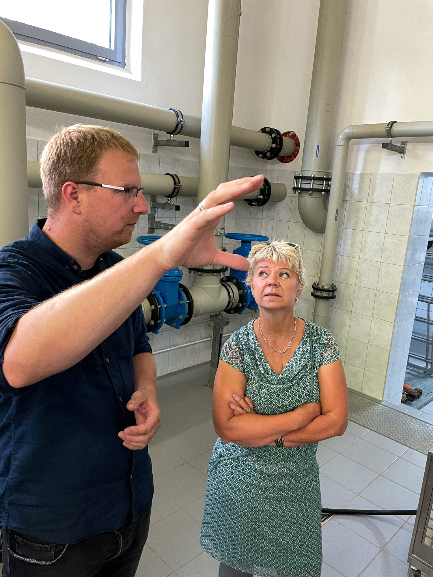 KI generiert: Das Bild zeigt zwei Personen in einem technischen oder industriellen Umfeld, die miteinander sprechen. Im Hintergrund sind Rohre und Armaturen zu sehen.