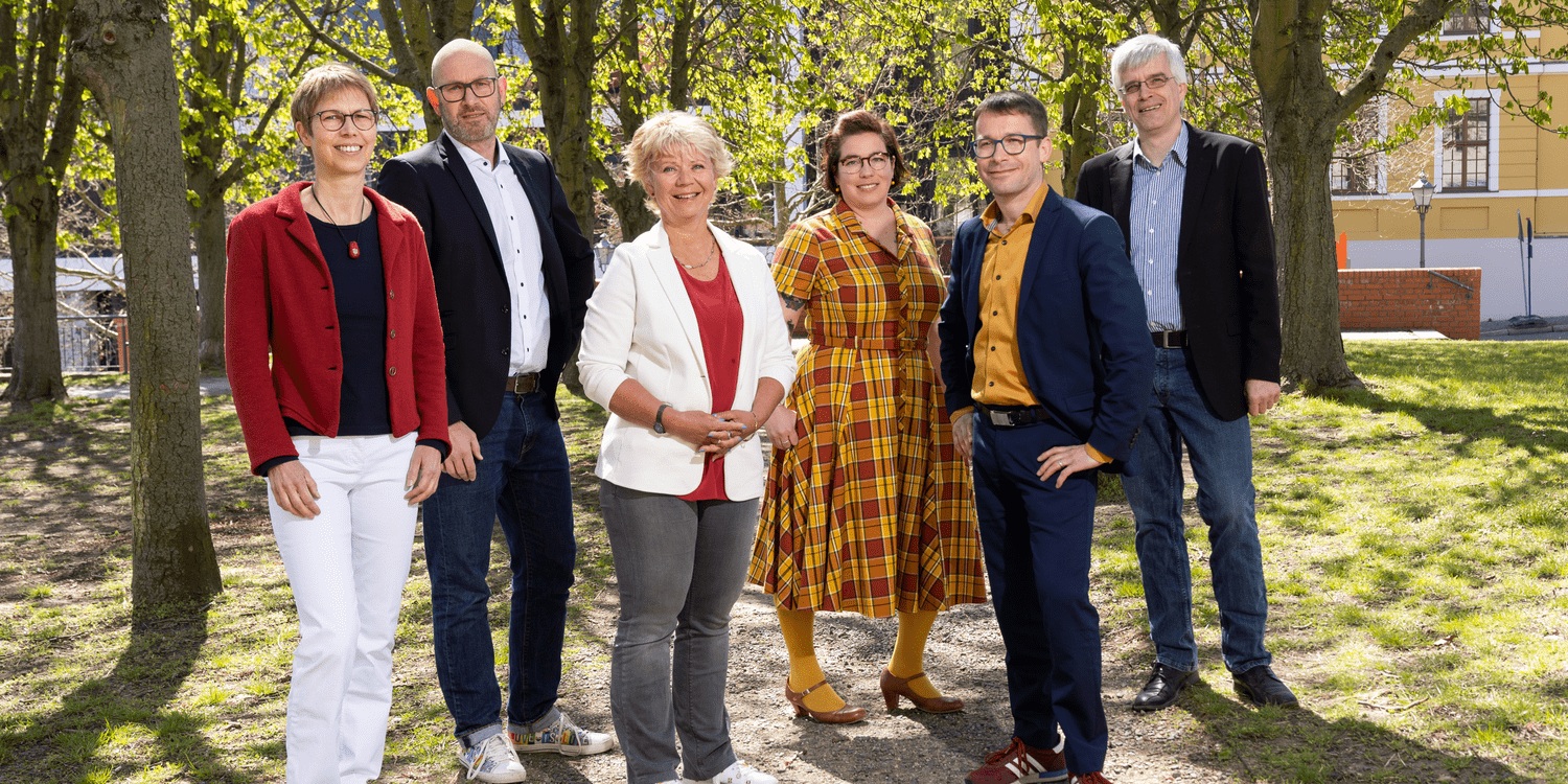 KI generiert: Eine Gruppe von sechs Personen steht lächelnd in einem sonnigen Park. Sie sind lässig-elegant gekleidet und posieren in einer freundlichen, offenen Umgebung.