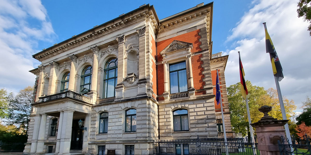 KI generiert: Das Bild zeigt ein historisches Gebäude mit einer aufwendigen Fassade und großen Fenstern. Vor dem Gebäude sind mehrere Flaggen aufgestellt.