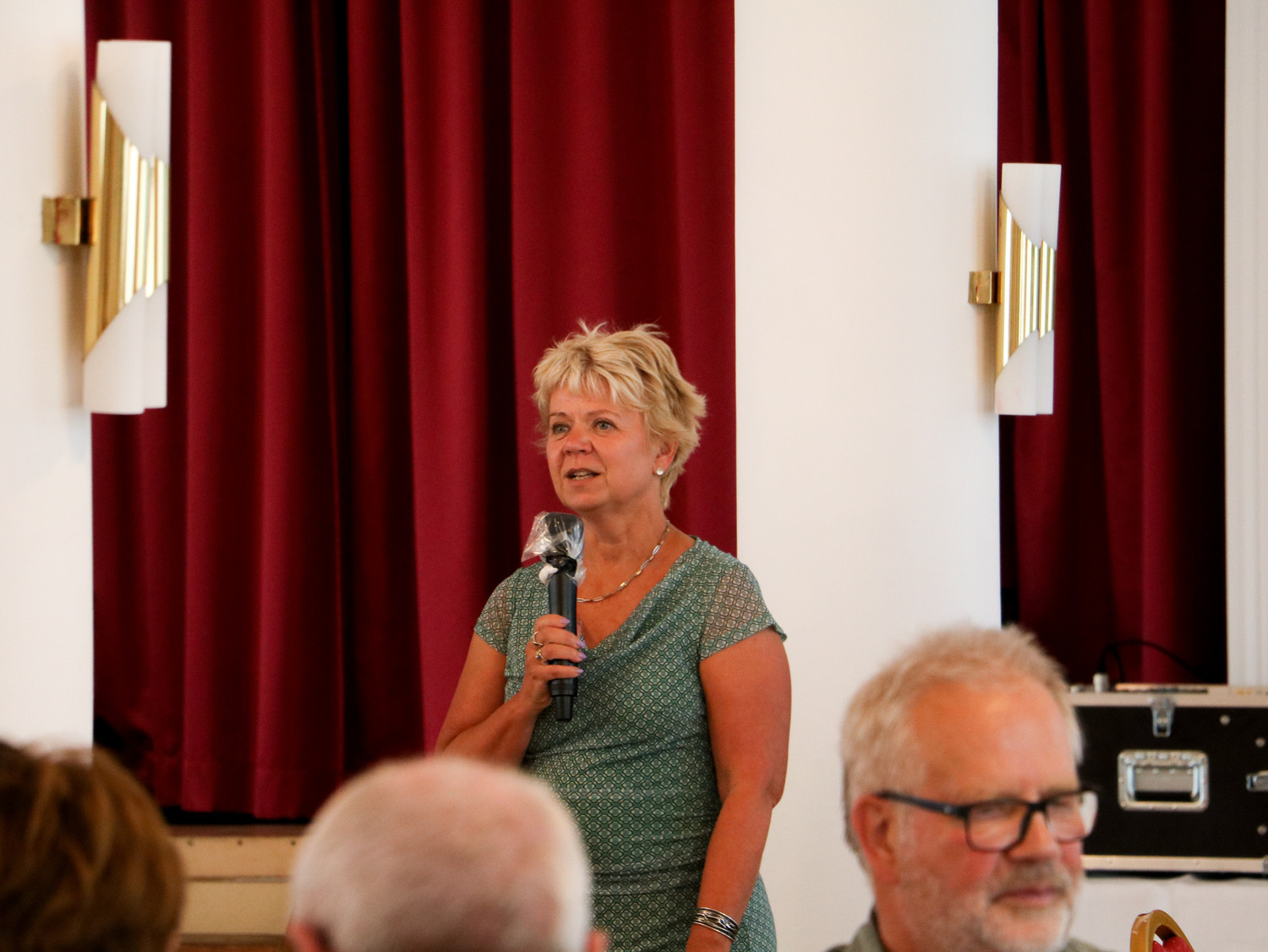 KI generiert: Eine Frau steht vor einem roten Vorhang und spricht in ein Mikrofon bei einer Veranstaltung. Im Vordergrund sind unscharf andere Personen zu sehen, die zuhören.