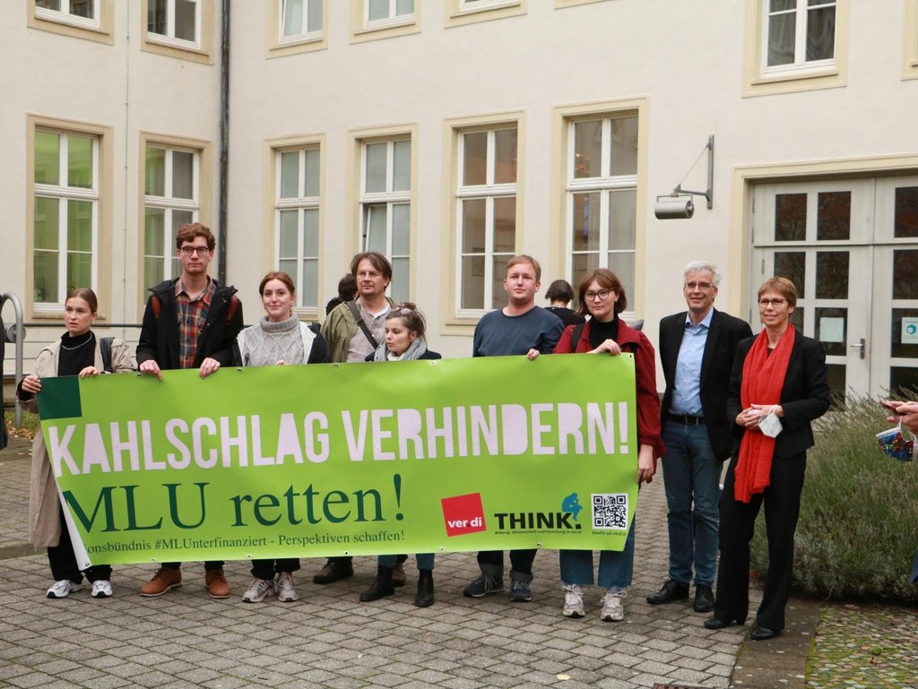 KI generiert: Eine Gruppe von Menschen steht vor einem Gebäude und hält ein grünes Banner mit der Aufschrift "Kahlschlag verhindern! MLU retten!". Das Banner weist auf eine Aktion hin, die sich gegen Kürzungen richtet.