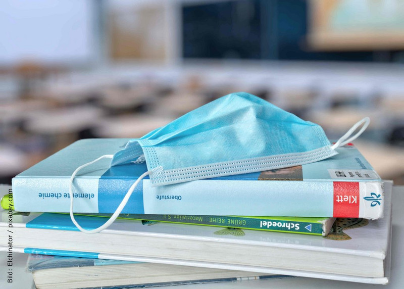 KI generiert: Das Bild zeigt einen Stapel Bücher auf einem Tisch, auf dem eine blaue medizinische Maske liegt. Im Hintergrund sind unscharf Schulmöbel und eine Tafel erkennbar.