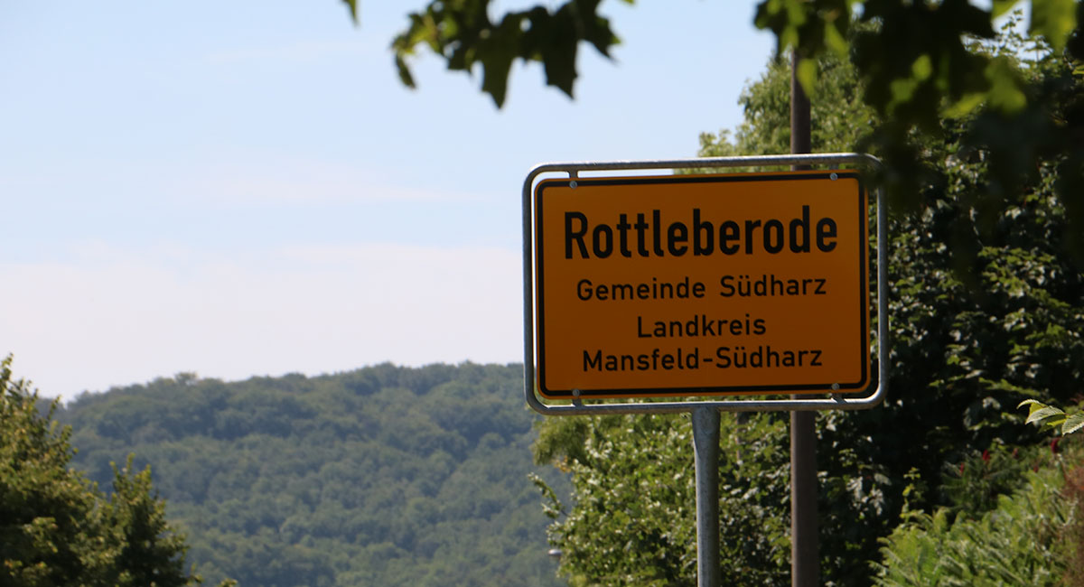 KI generiert: Das Bild zeigt ein Ortsschild von Rottleberode in der Gemeinde Südharz, Landkreis Mansfeld-Südharz. Im Hintergrund sind Bäume und Hügel zu sehen.
