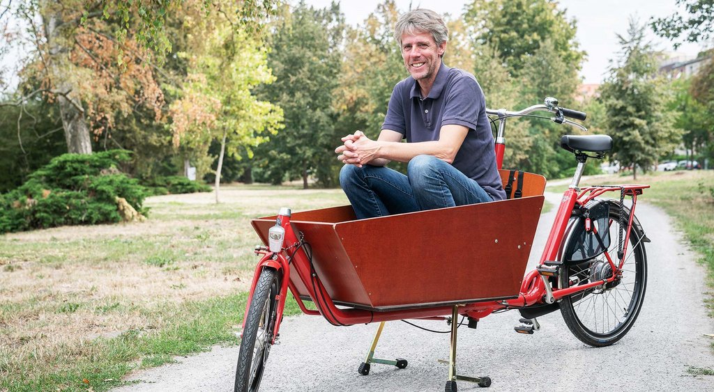 Stephan Marahrens hockt in der Transportbox eines roten Lastenfahrrades und lächelt in die Kamera. Sie befinden sich auf einem Schotterweg in einem Park. 