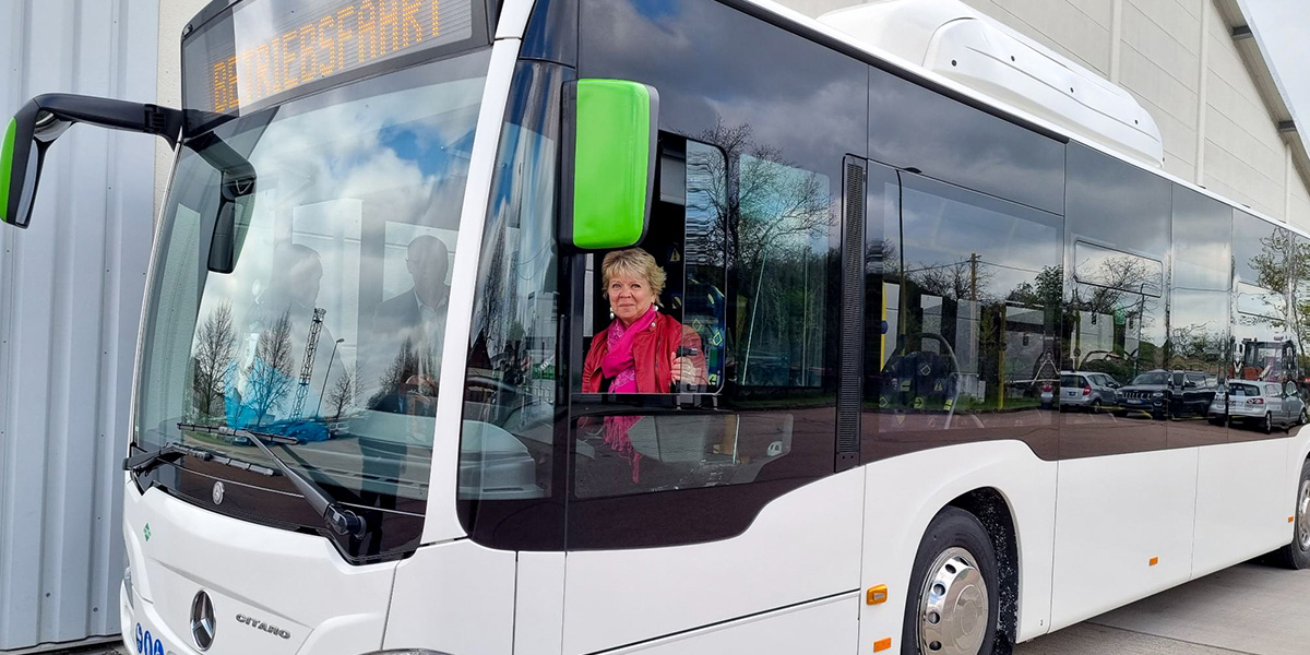 KI generiert: Das Bild zeigt einen modernen Bus, der vor einem Gebäude geparkt ist, mit einer Person, die im Führerhaus sitzt. Die Anzeigetafel des Busses zeigt den Schriftzug "ÖPNV Erprobung".