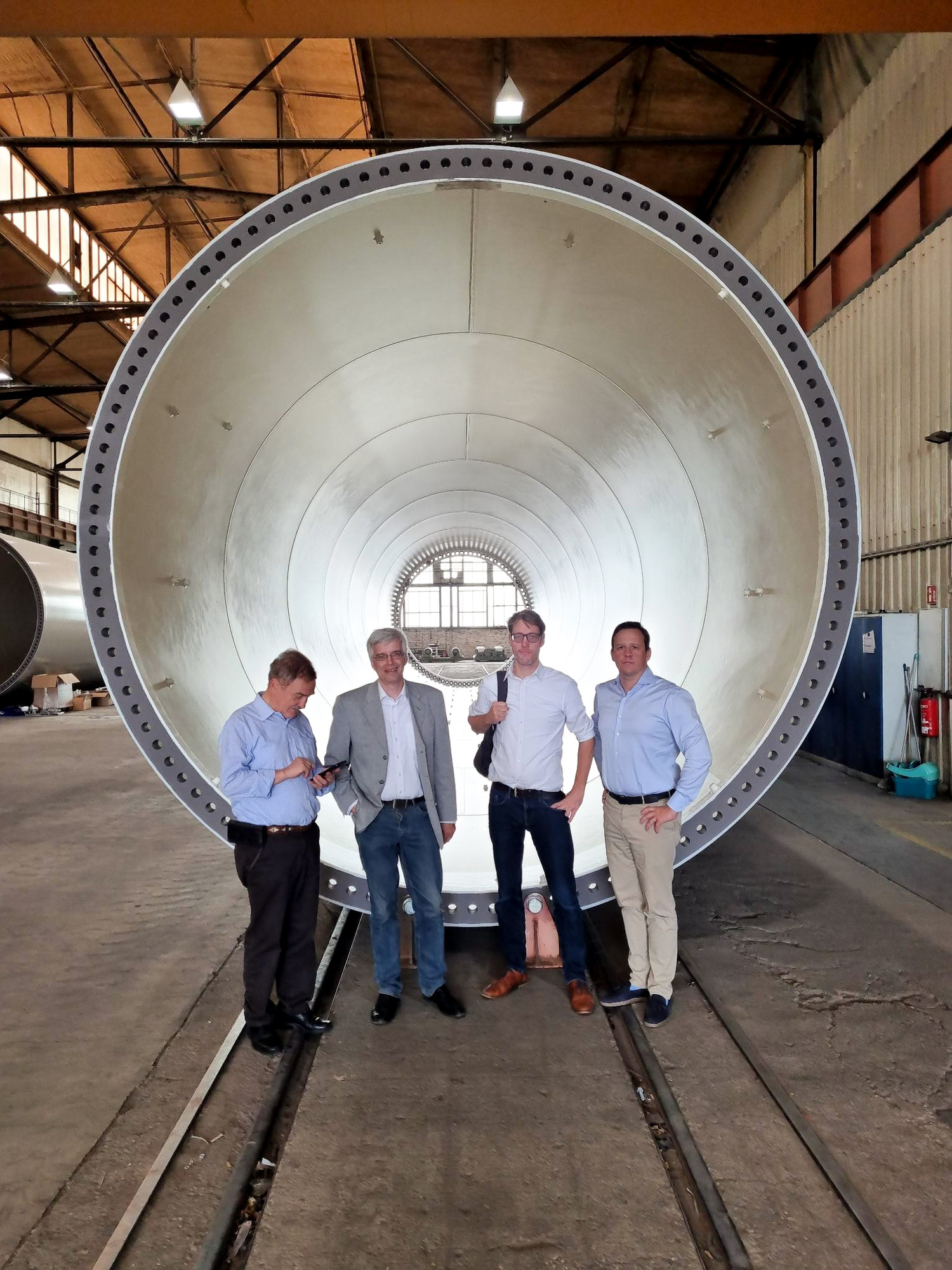KI generiert: Das Bild zeigt vier Männer, die vor einem großen, zylindrischen Metallbauteil in einer Werkhalle stehen. Sie stehen auf einem Schienensystem innerhalb der Halle.