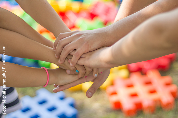 KI generiert: Auf dem Bild legen mehrere Menschen ihre Hände übereinander, was Zusammenhalt und Teamarbeit symbolisiert. Im Hintergrund sind bunte Bausteine zu sehen.