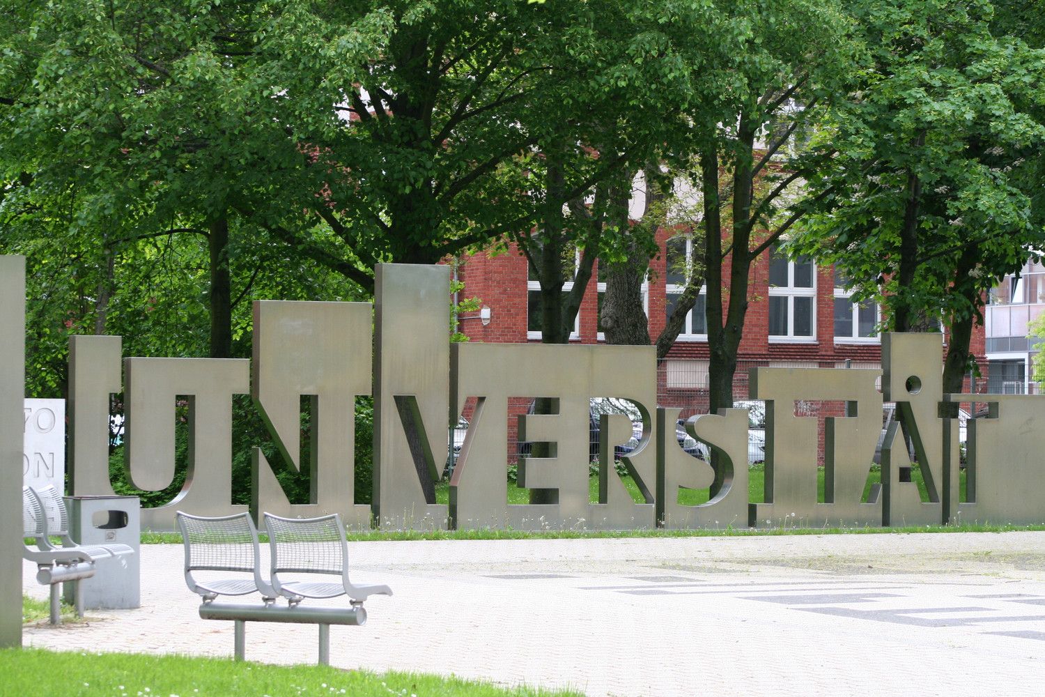 KI generiert: Das Bild zeigt eine große Skulptur mit dem Wort "UNIVERSITÄT" in einem grünen, parkähnlichen Bereich. Im Hintergrund sind Bäume und ein Backsteingebäude sichtbar.