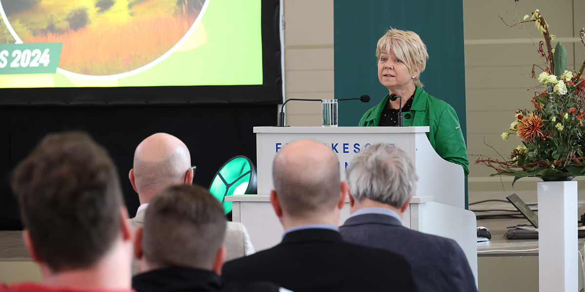 KI generiert: Das Bild zeigt eine Frau, die an einem Podium steht und vor einer Gruppe von Menschen spricht. Im Hintergrund ist eine Leinwand mit einer Präsentation und Dekorationen zu sehen.
