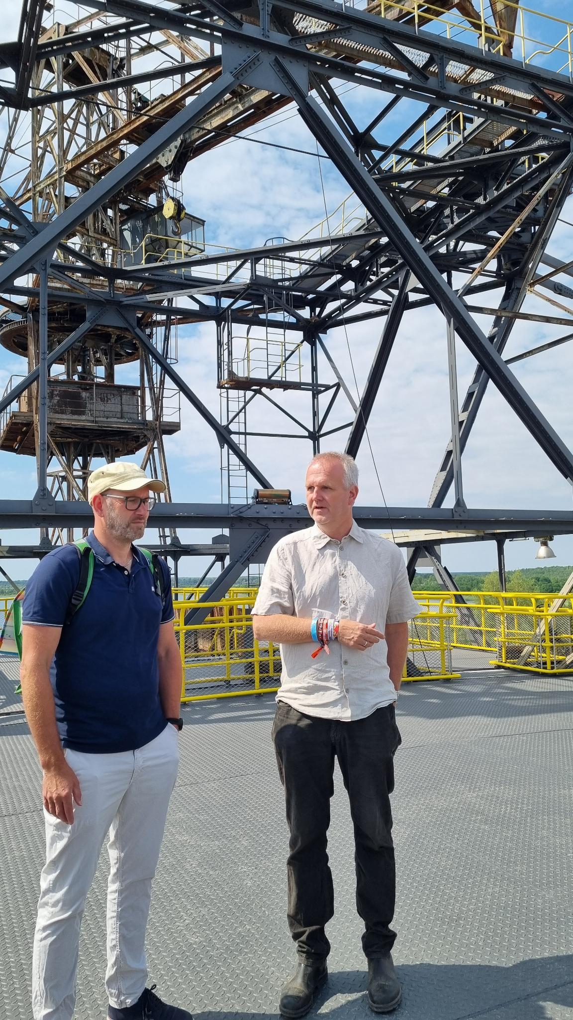KI generiert: Das Bild zeigt zwei Männer vor einer großen Metallstruktur, die vermutlich Teil eines industriellen Komplexes oder Förderanlage ist. Die Männer stehen auf einer Plattform und scheinen sich zu unterhalten.