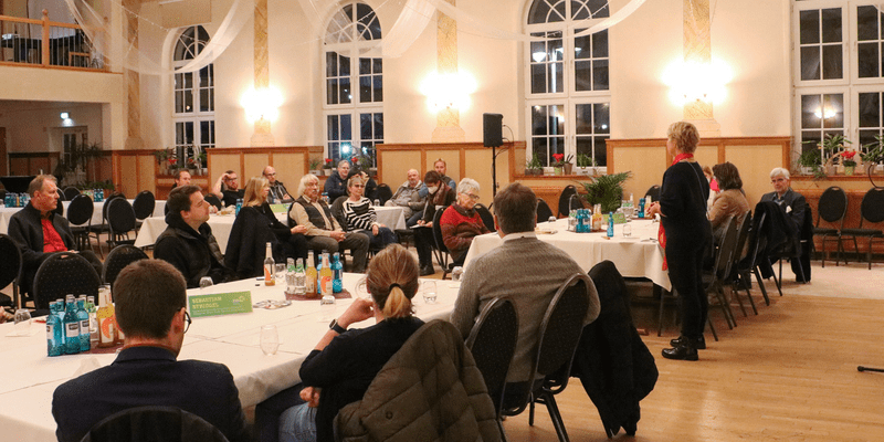 KI generiert: Das Bild zeigt eine Versammlung oder Diskussion in einem Gemeinschaftsraum, bei der ein stehender Redner oder Moderator vor einem sitzenden Publikum spricht. Der Raum ist mit runden Tischen und Stühlen arrangiert.