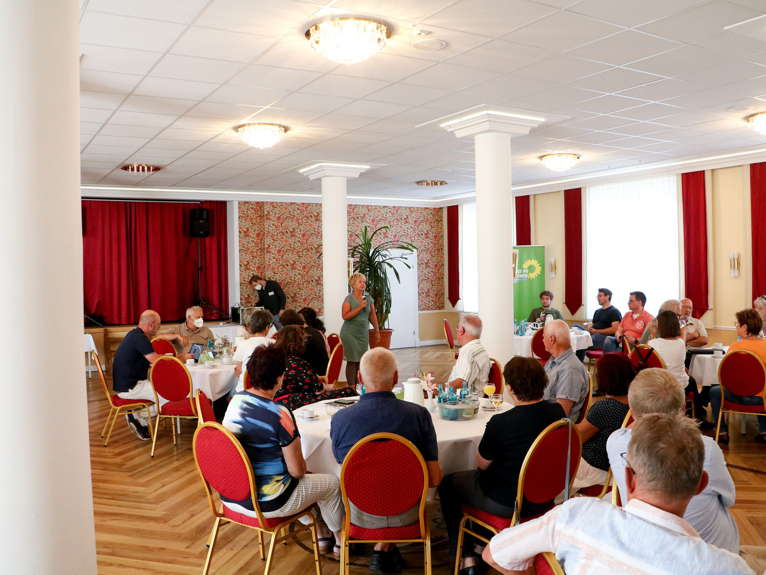 KI generiert: Eine Gruppe von Menschen sitzt in einem Veranstaltungsraum an Tischen, während eine Person vorne zu sprechen scheint. Im Hintergrund ist ein Banner mit einem grünen Motiv zu sehen.