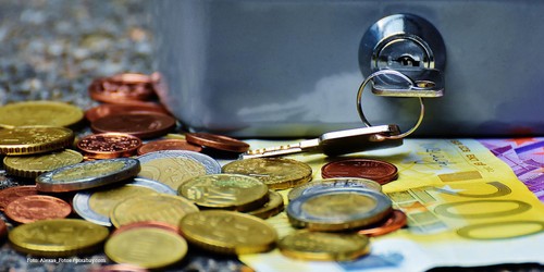 KI generiert: Auf dem Bild liegen mehrere Euro-Münzen und ein 200-Euro-Schein vor einem geschlossenen Metallkästchen, in dessen Schloss ein Schlüssel steckt. Der Hauptinhalt des Bildes ist Geld in Form von Münzen und einem Schein.