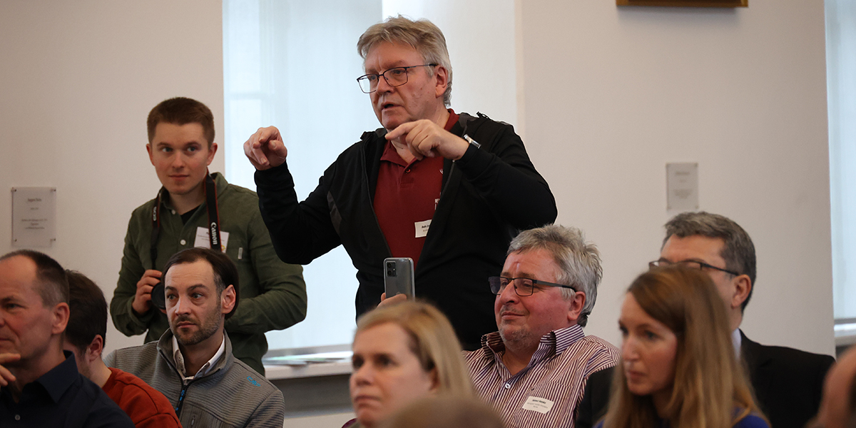 KI generiert: Das Bild zeigt eine Gruppe von Menschen in einem Raum, von denen einer steht und offenbar etwas erklärt oder eine Frage stellt. Die Anwesenden scheinen einer Diskussion oder Präsentation beizuwohnen.