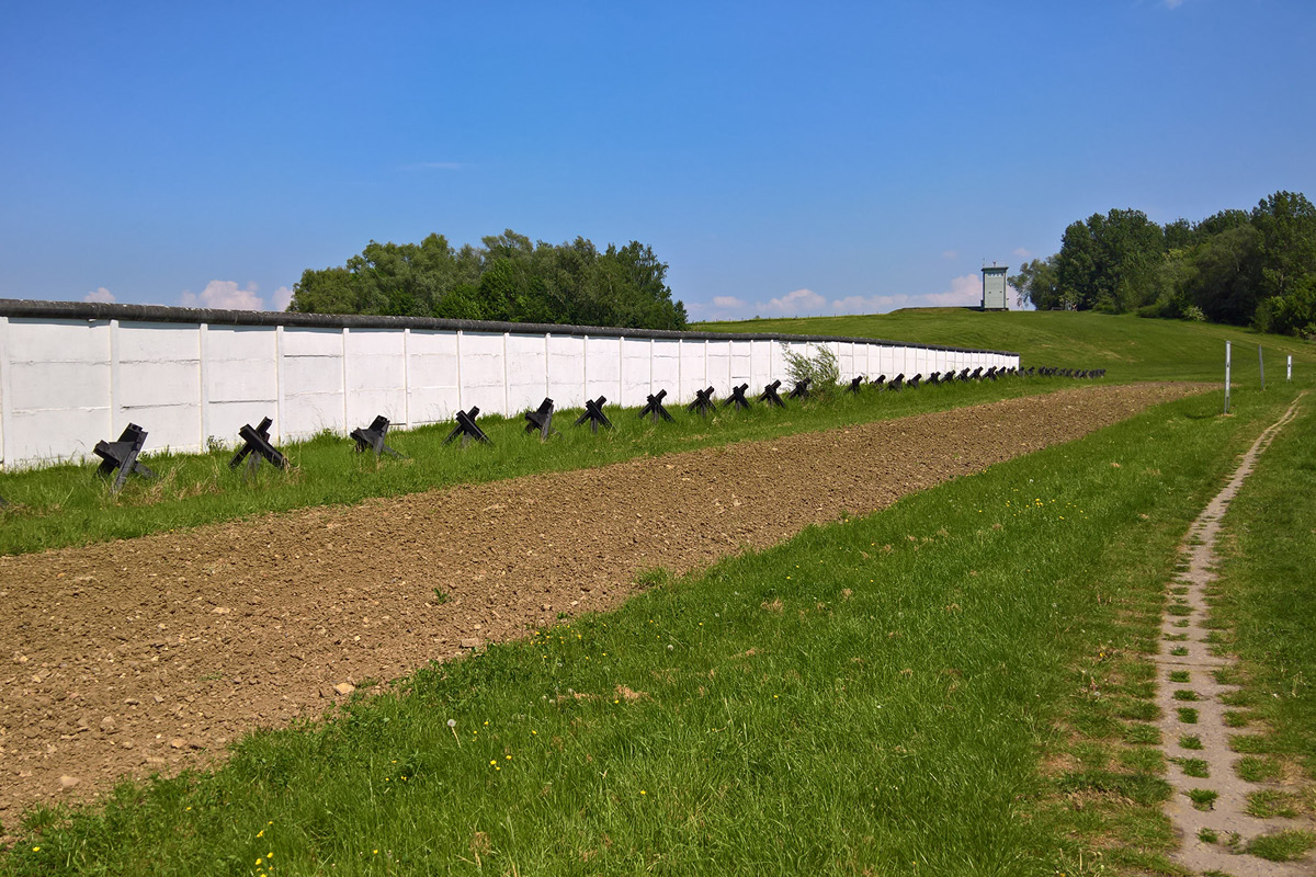 KI generiert: Das Bild zeigt eine lange weiße Mauer mit Verteidigungsstrukturen davor, die an einen Abschnitt der Berliner Mauer erinnern. Daneben verläuft ein schmaler Pfad durch eine grüne Wiese.