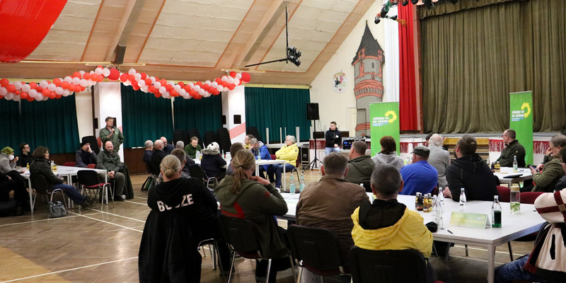 KI generiert: Das Bild zeigt eine Versammlung oder Diskussionsrunde in einem Saal mit Menschen, die um Tische herum sitzen, während eine Person vorne spricht. Grüne Banner deuten auf eine Veranstaltung im Zusammenhang mit den Grünen oder einer ähnlich ausgerichteten Organisation hin.