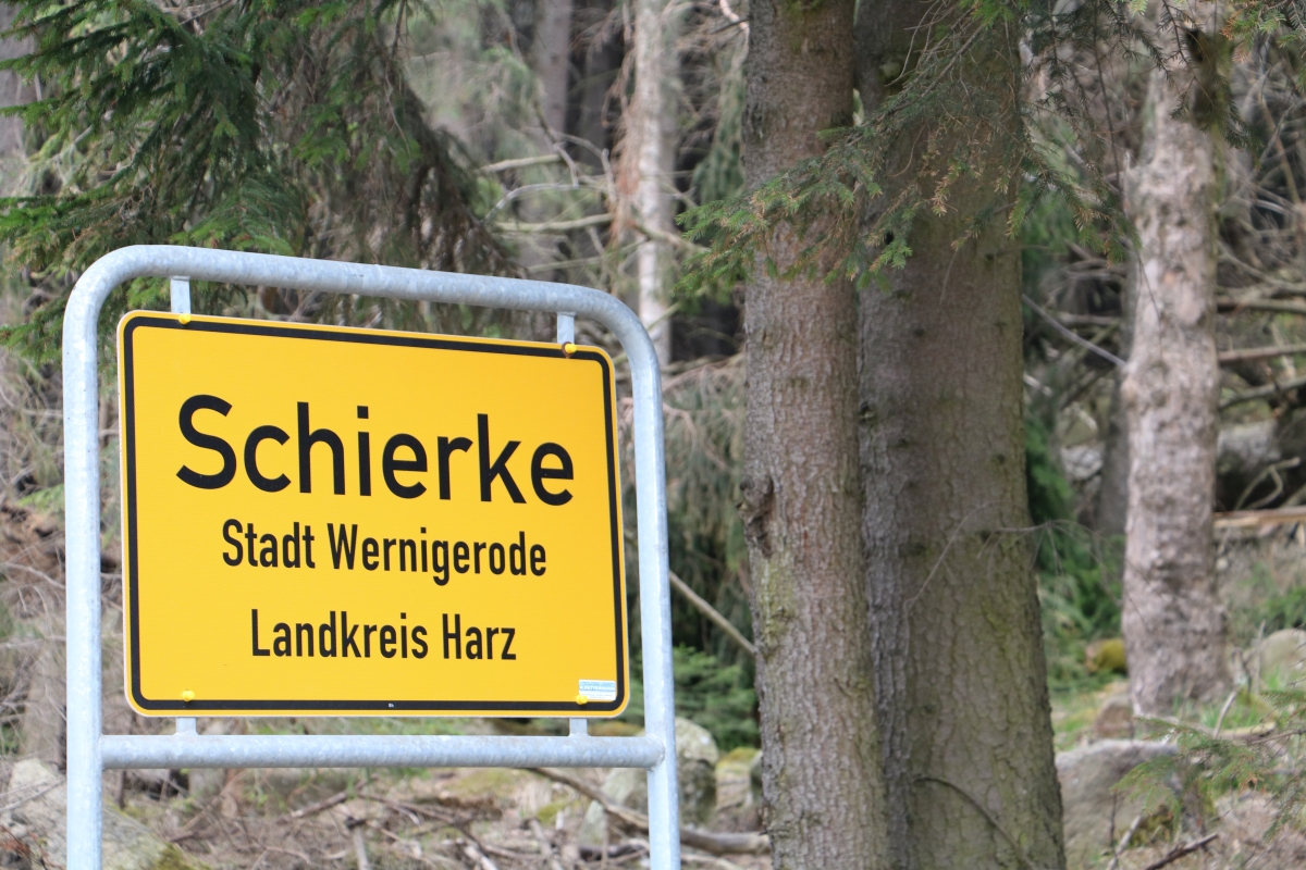 KI generiert: Das Bild zeigt ein gelbes Ortsschild mit der Aufschrift "Schierke", das zur Stadt Wernigerode im Landkreis Harz gehört. Im Hintergrund sind Bäume zu sehen, die auf eine waldreiche Gegend hindeuten.