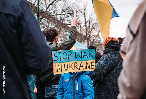 KI generiert: Das Bild zeigt Menschen auf einer Demonstration, die ein Schild mit der Aufschrift "STOP WAR IN UKRAINE" tragen. Im Hintergrund sind eine ukrainische Flagge und weitere Personen zu sehen.