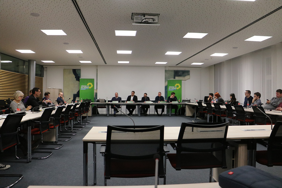 KI generiert: Das Bild zeigt eine Besprechung oder Konferenz in einem Raum mit Teilnehmern, die um einen U-förmigen Tisch sitzen. Im Hintergrund sind Banner mit einem grünen Logo zu sehen.